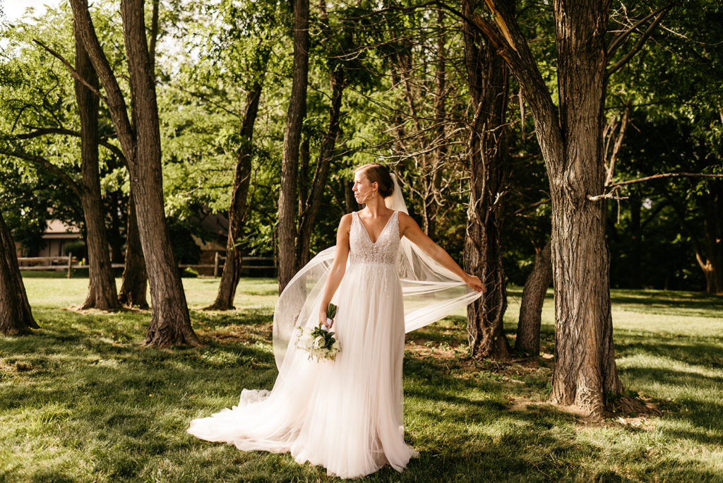 bride in a luxury gown standing in the woods with her veil blowing in the wind behind her as she holds her floral wedding bouquet
