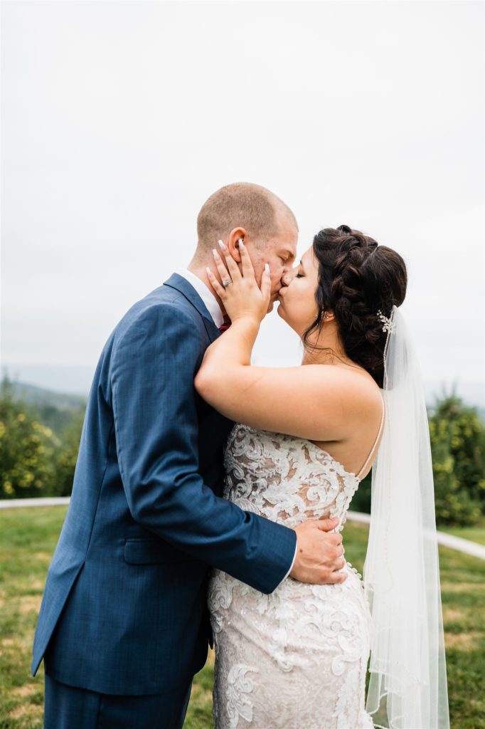 Shenandoah wedding with Charlottesville wedding photographer captures bride and groom kissing on a hill top as they embrace each other with bride in a lace wedding dress and groom in a blue suit