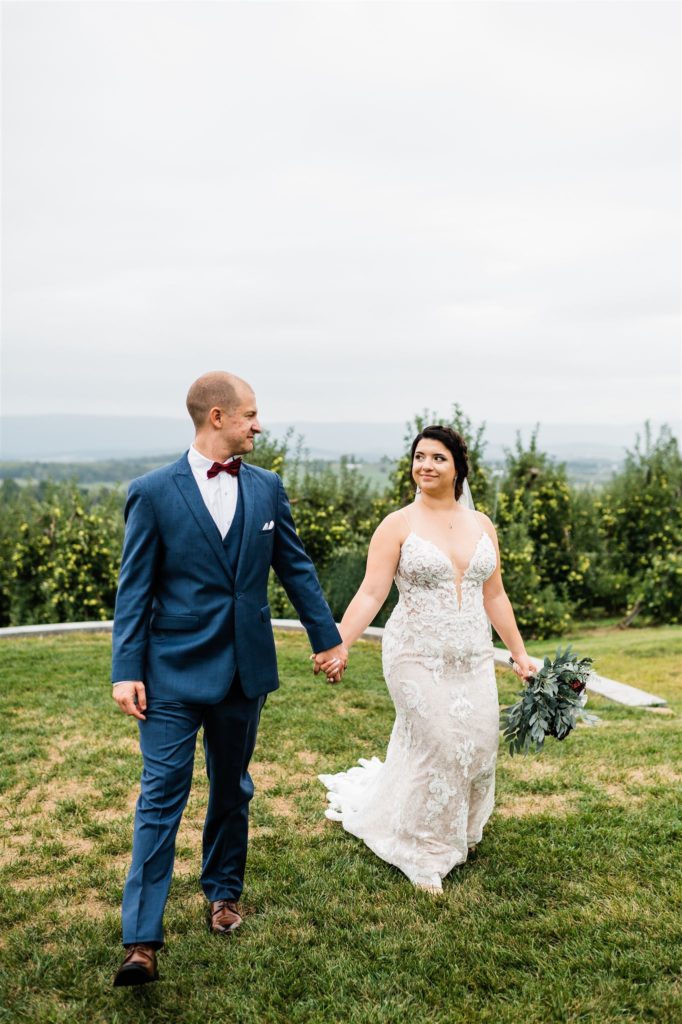 Charlottesville wedding photographer photographs bride and groom holding hands and walking on a hill with an orchard behind them