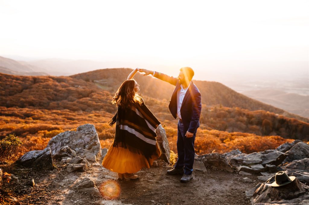 Shenandoah engagement pictures with Charlottesville wedding photographer with man and woman on a mountain top dancing while the sun shines in the distance as it sets over the trees and mountains 