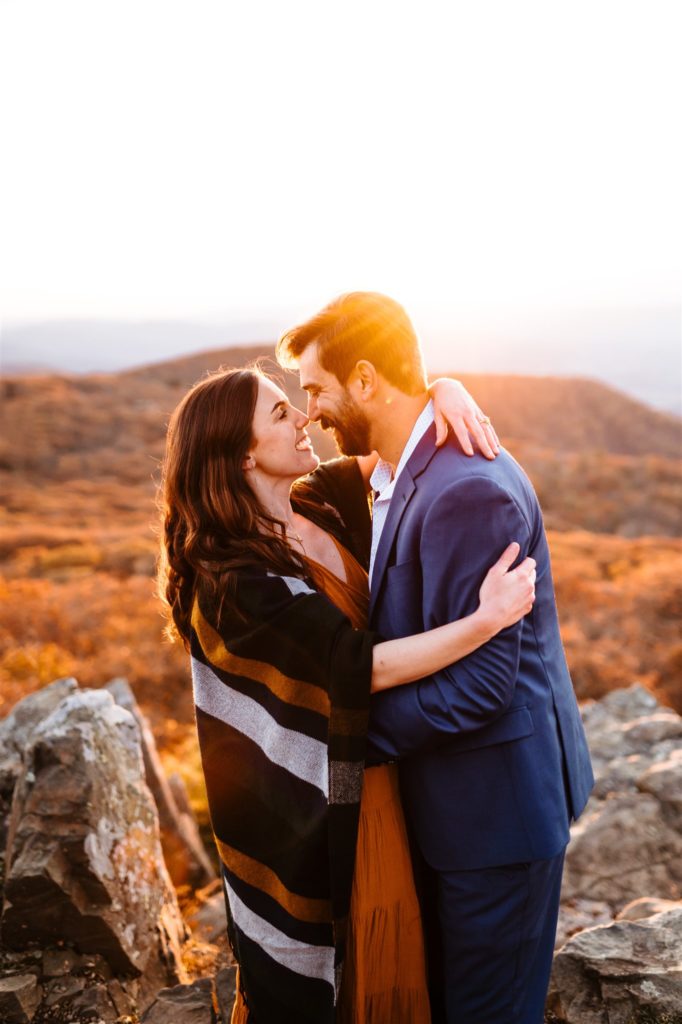 Charlottesville wedding photographer captures engagement pictures at sunset in Shenandoah with man and woman embracing on a mountain top while the sun sets in the distance