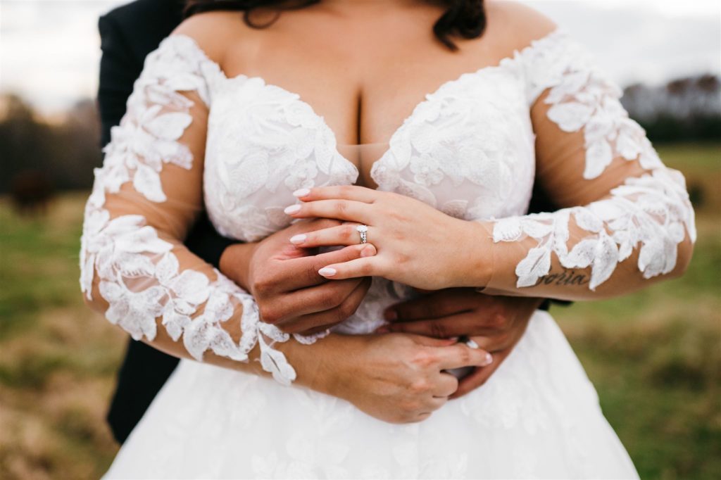 detail shot of brides engagement ring and wedding ring as she holds her grooms hands while he wraps his arms around her waist taken by Charlottesville wedding photographer