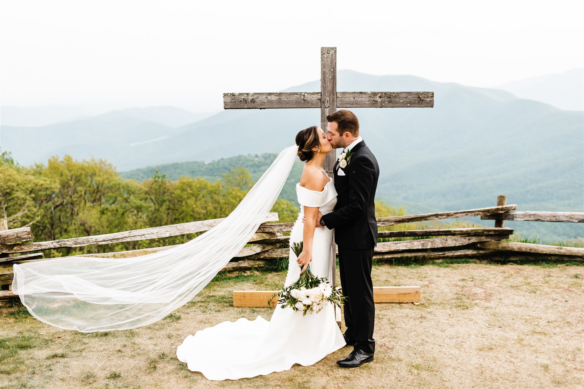 Shenandoah National Park Wedding with bride and groom kissing next to a wooden cross on a mountain top with brides veil flowing in the wind behind her