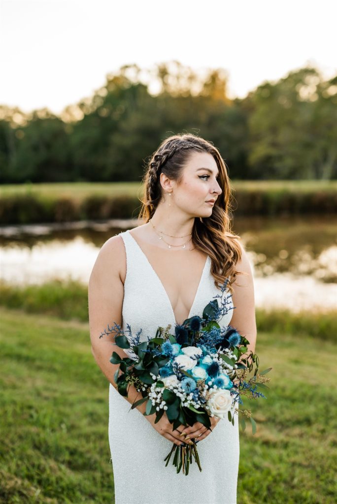 bride in a satin and embellished wedding dress holding a blue floral wedding bouquet and looking off the the distance stands in front of a lake captured by Charlottesville wedding photographer