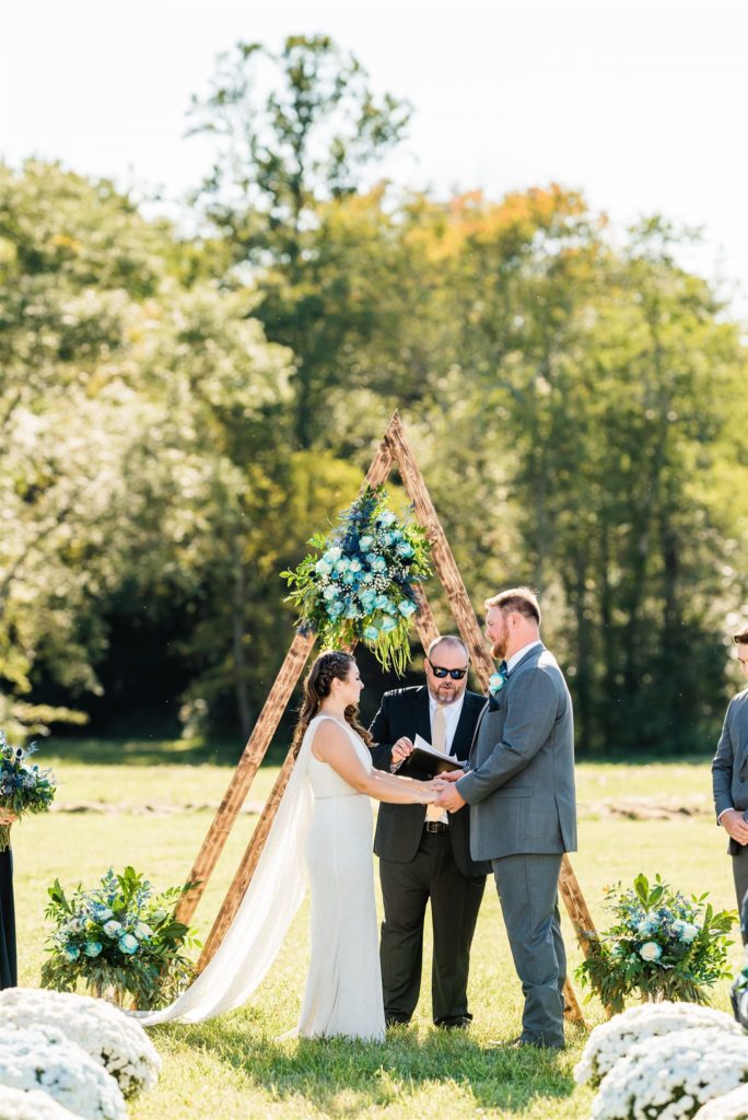 bride and groom holding hands during their outdoor ceremony with a triangle arch decorated with blue florals captured by Charlottesville wedding photographer 