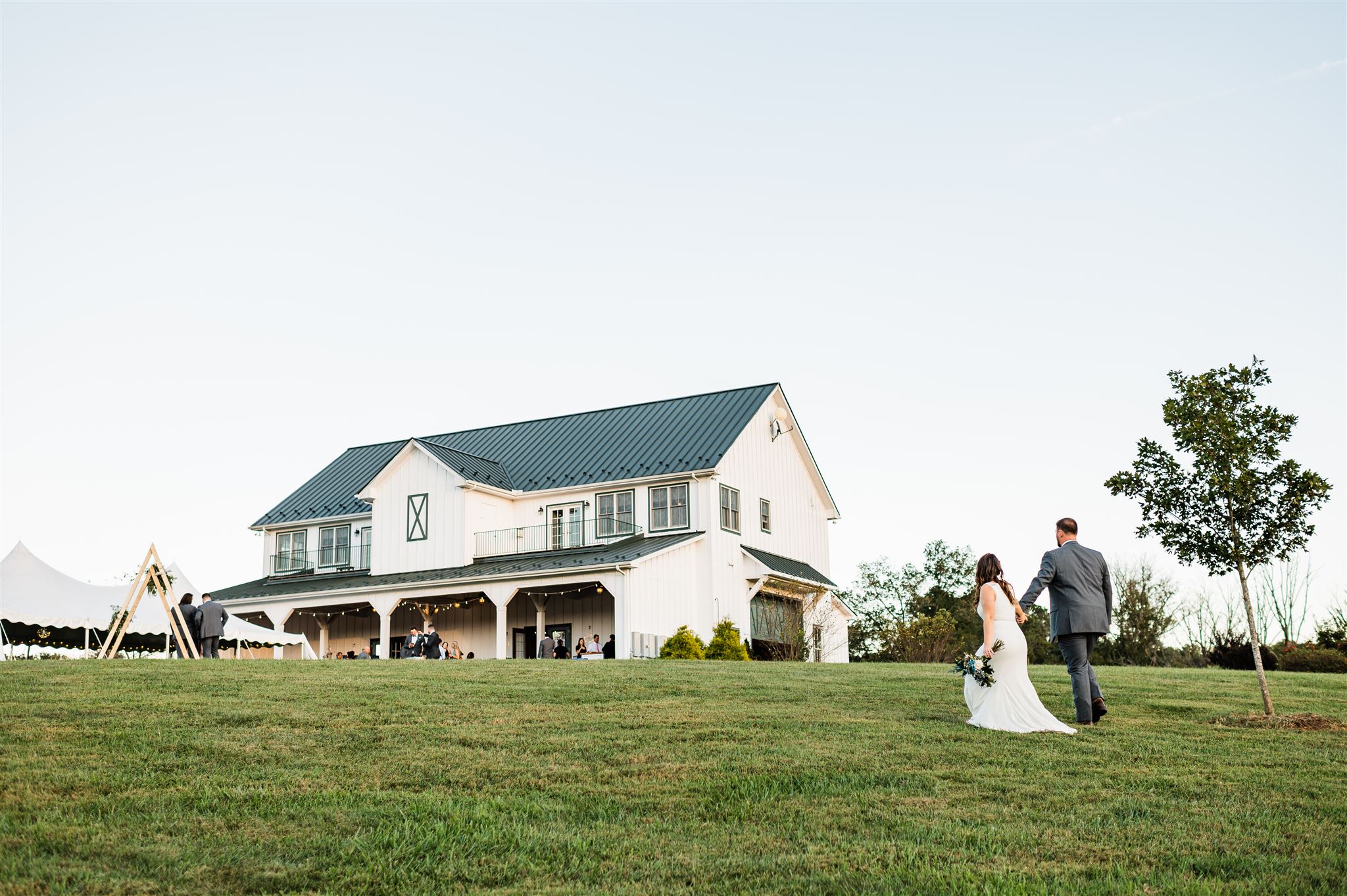 bride and groom holding hands and walking up a hill together at their wedding venue in Shenandoah with the white barn wedding venue in the distance