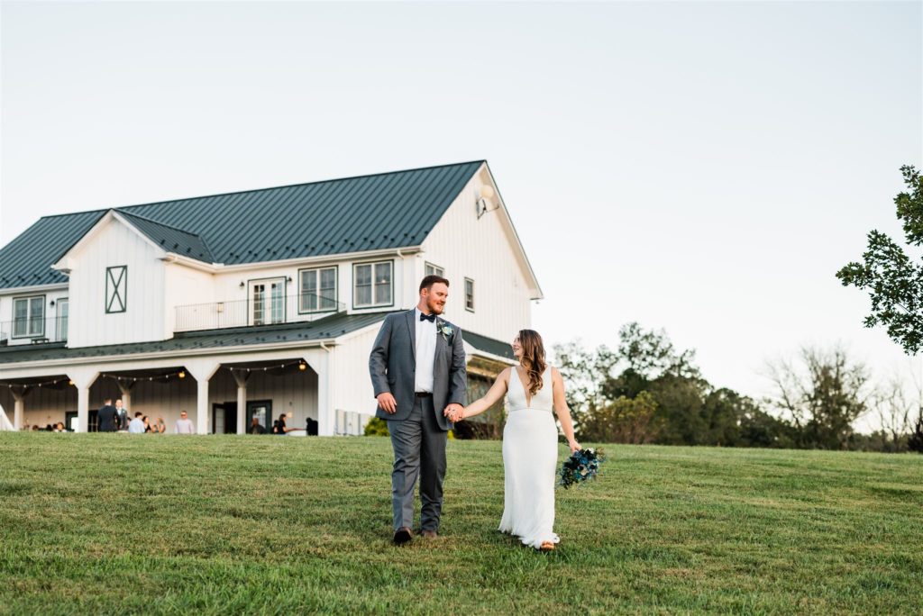 Charlottesville wedding photographer captures bride and groom holding hands and walking down a hill with their Shenandoah wedding  venue in the distance while they look to each other and smile 