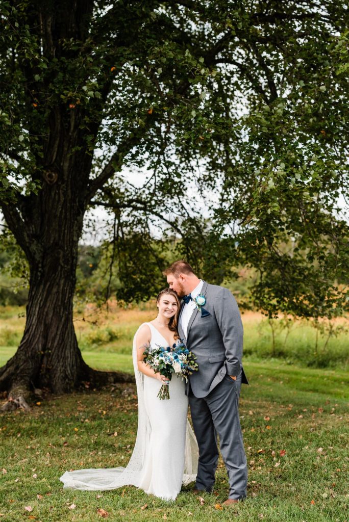 bride and groom standing under a tree at their Charlottesville wedding venue as the bride holds a blue floral bouquet and the groom leans down to kiss her 