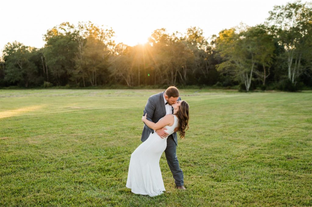 Shenandoah wedding pictures at sunset with groom dipping his bride backwards while kissing her in a field with trees in the distance photographed by Charlottesville wedding photographers