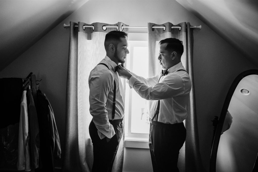 groom with his father getting ready for the wedding day as the father helps his son with his bow tie in front of a window 