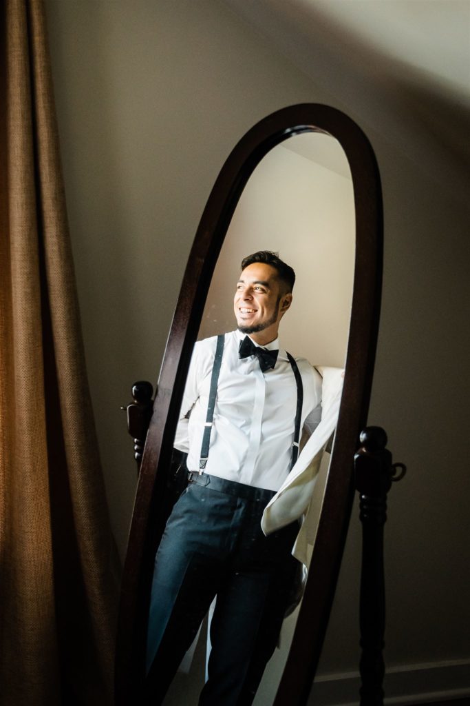 Shenandoah wedding with groom getting ready before his wedding day while standing in a tall mirror next to a window