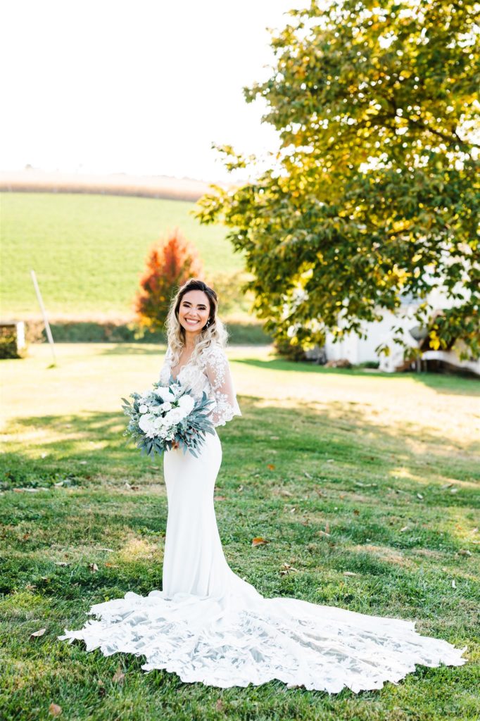 Charlottesville wedding photographer captures bride in a lace wedding dress with a large train standing on the lawn of her Shenandoah wedding venue while holding her white rose bouquet and smiling 