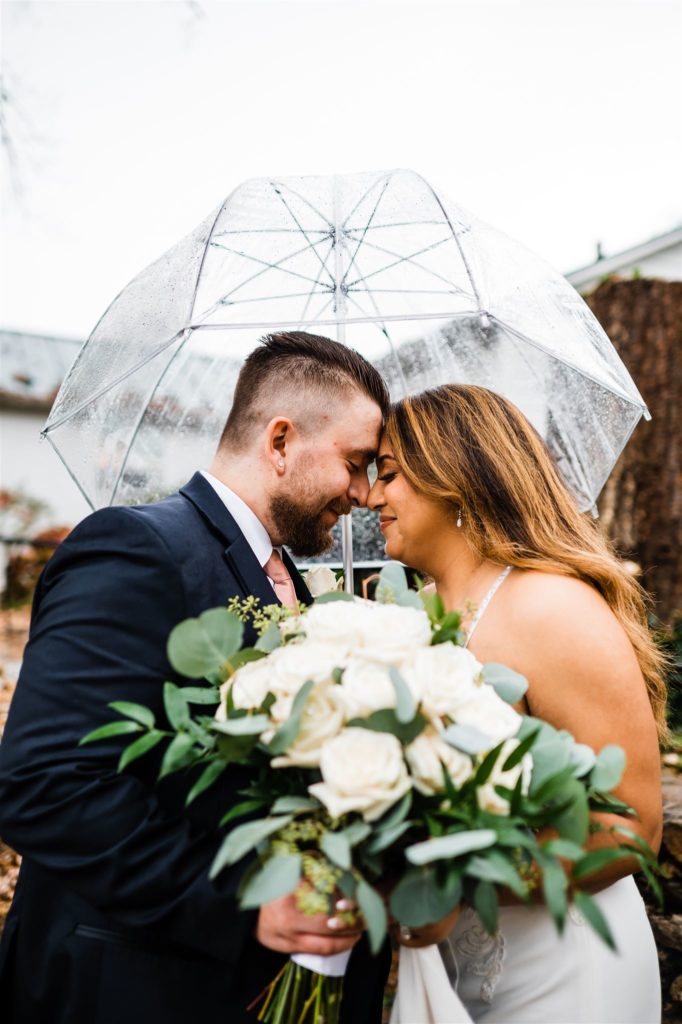 rainy wedding day photos with bride and groom standing under a clear umbrella with the brides white rose bouquet