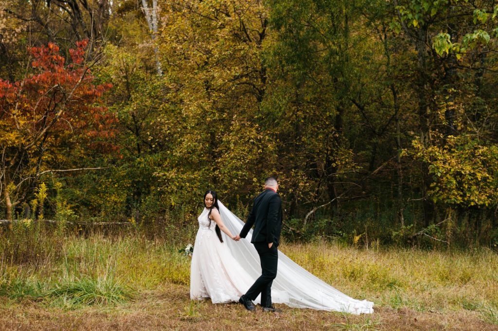 Charlottesville wedding photographer captured bride and groom holding hands and dancing in a field  with a near by grove of trees in the fall for a Shenandoah wedding 