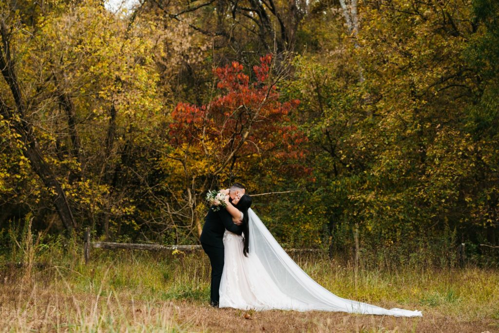 fall Shenandoah wedding with bride and groom in a field next to a line of trees filled with fall colors embracing romantically and kissing captured by Charlottesville wedding photographer 