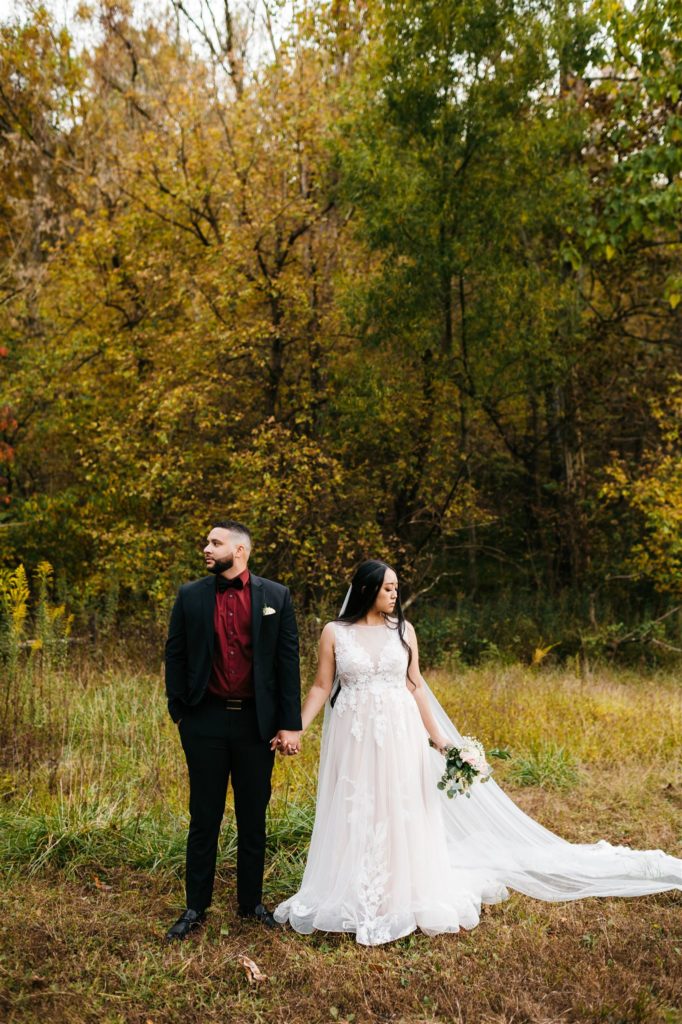 Charlottesville wedding photographer captures bride and groom holding hands and looking opposite directions in a field with a tree line in the distance for their Shenandoah wedding 