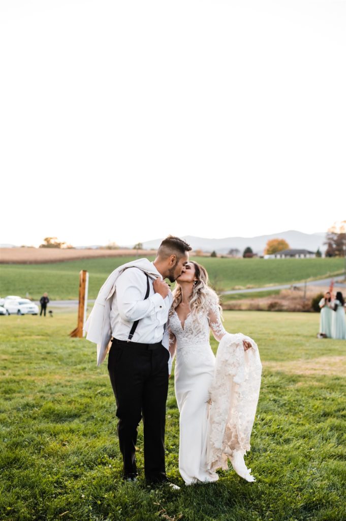 Charlottesville wedding photographer captures bride and groom holding hands and kissing as they walk up a hill at their Shenandoah wedding venue while groom holds his suit coat over his shoulder and the bride holds her dress train 
