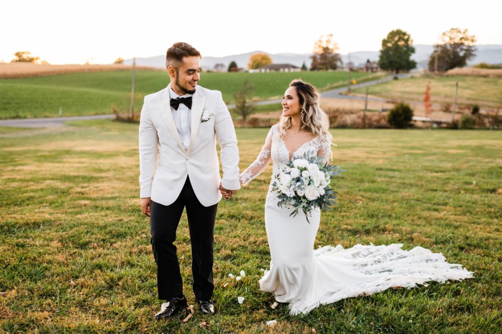 Charlottesville wedding photographer captures bride and groom holding hands and walking up a hill together as they look at one another with a farm  in the distance with rolling hills and mountains 