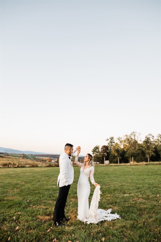Charlottesville wedding photographer captured Shenandoah wedding with bride and groom dancing at sunset in a large field with a row of trees in the distance and mountains even further in the distance 
