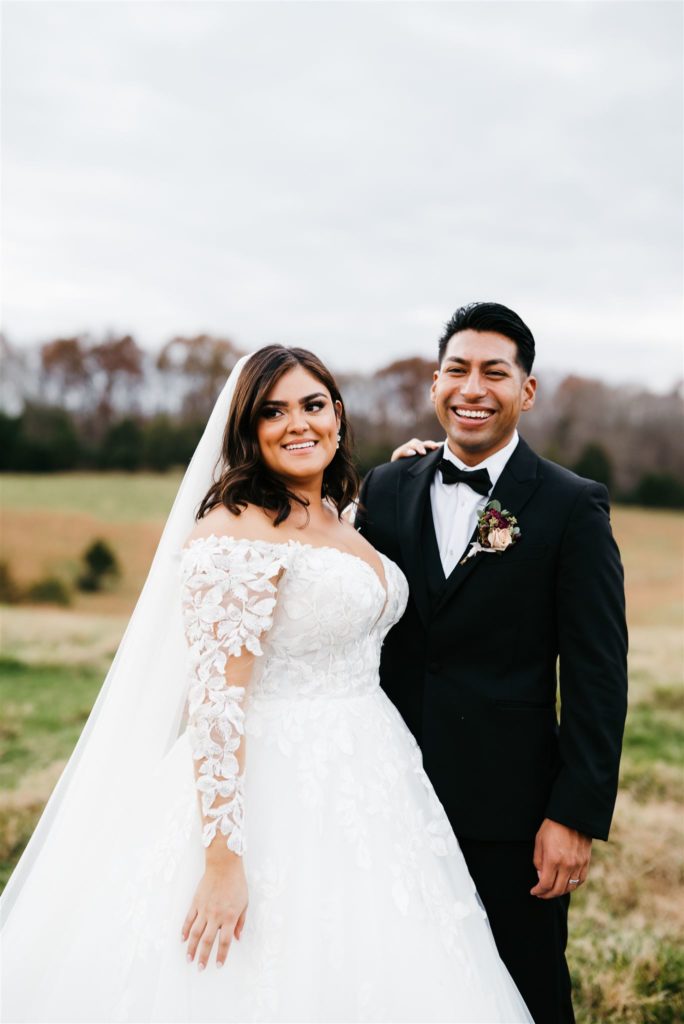 Charlottesville wedding photographer captures bride and groom standing together and smiling in a pasture with cows in the distance for their Shenandoah wedding on a cloudy day  