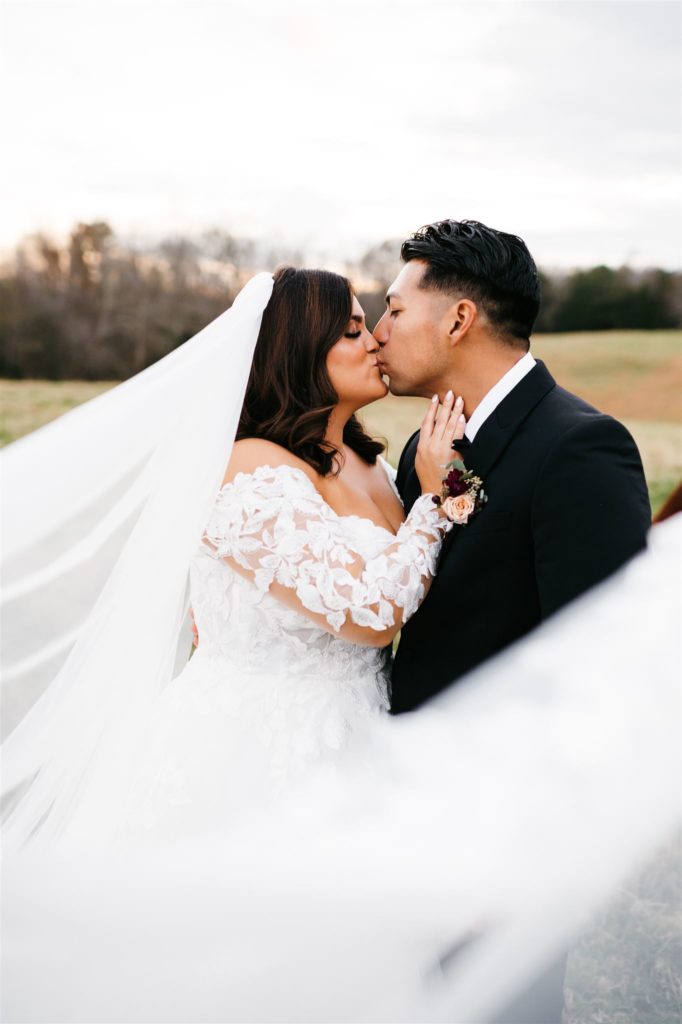 bride and groom kissing in a pasture at a Shenandoah wedding venue with brides veil blowing in the wind captured by Charlottesville wedding photographer