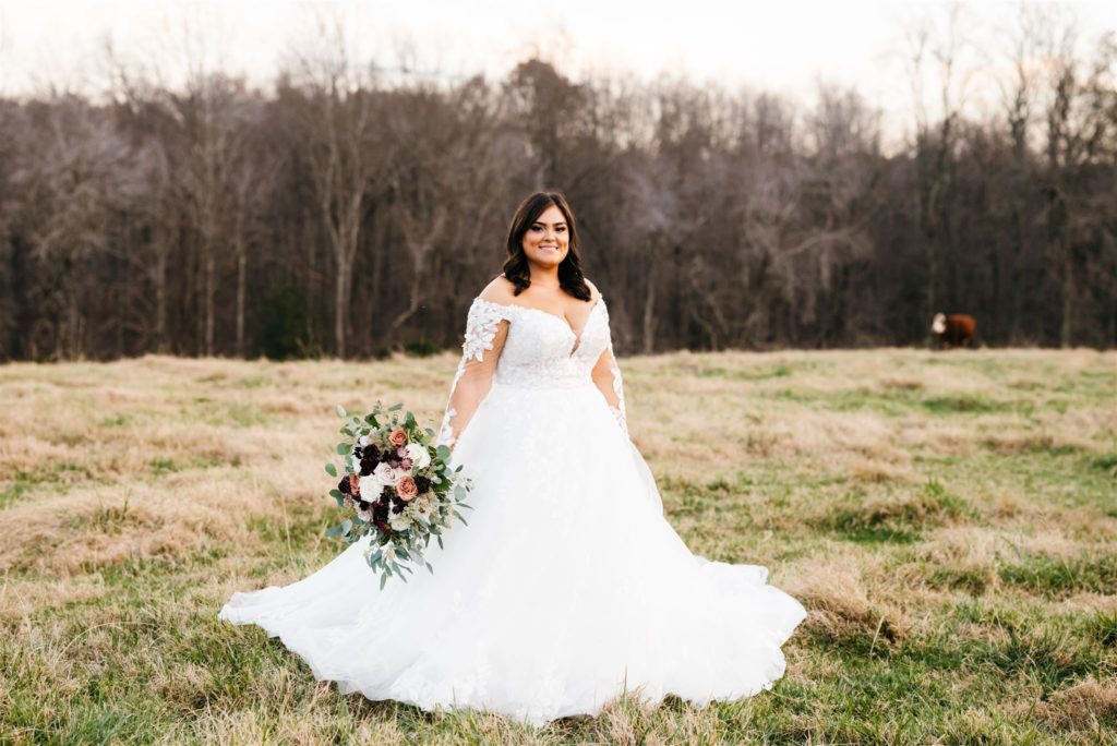 bride in a large lace and tulle wedding dress standing in a pasture for her Shenandoah wedding  holding a large burgundy and white wedding bouquet captured by Charlottesville wedding photographer 