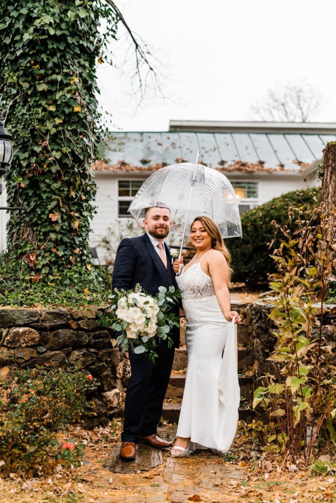 bride and groom in a garden with a stone wall embracing one another under a clear umbrella for their Shenandoah valley wedding venues wedding 
