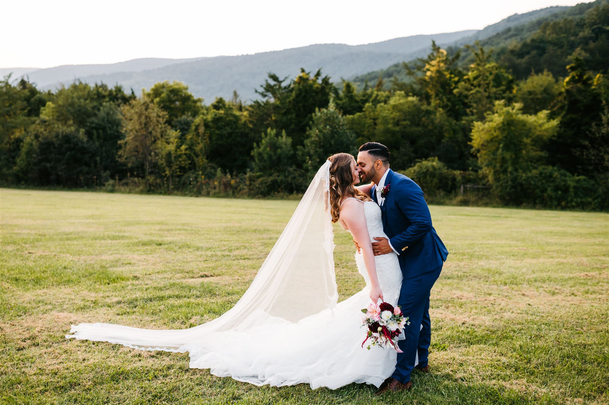 Shenandoah wedding with bride and groom in an open field with a sec tree line of evergreen forest behind them and mountains in the distance while the groom hold his bride by the waist and leans into her to kiss her passionately as she holds her wedding bouquet to her side captured by Virginia wedding photographer
