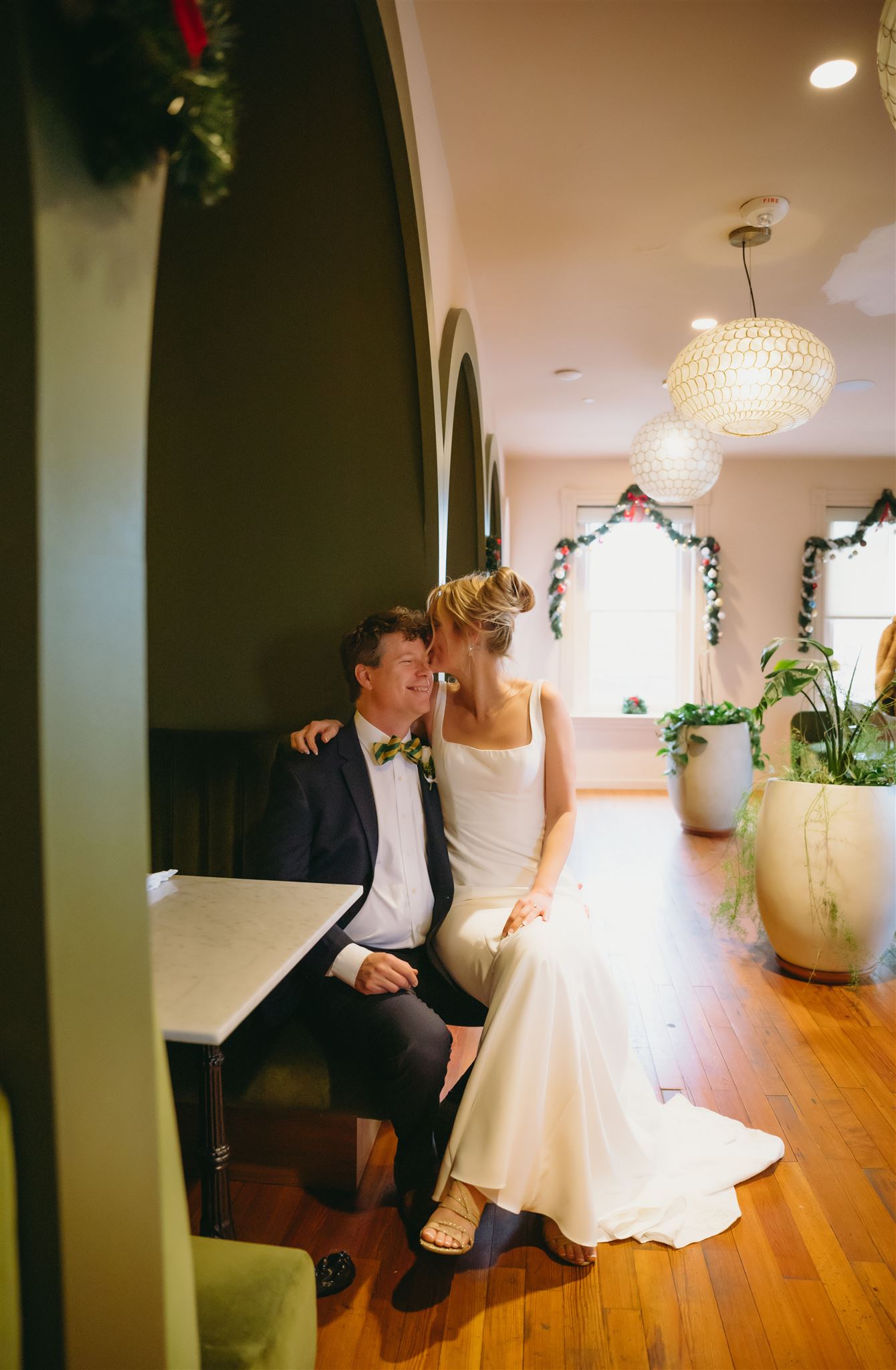 groom sitting in a booth at a cafe in a richmond wedding venue with his bride on his lap and kissing his head with holiday decor in the distance photographed by Richmond wedding photographer