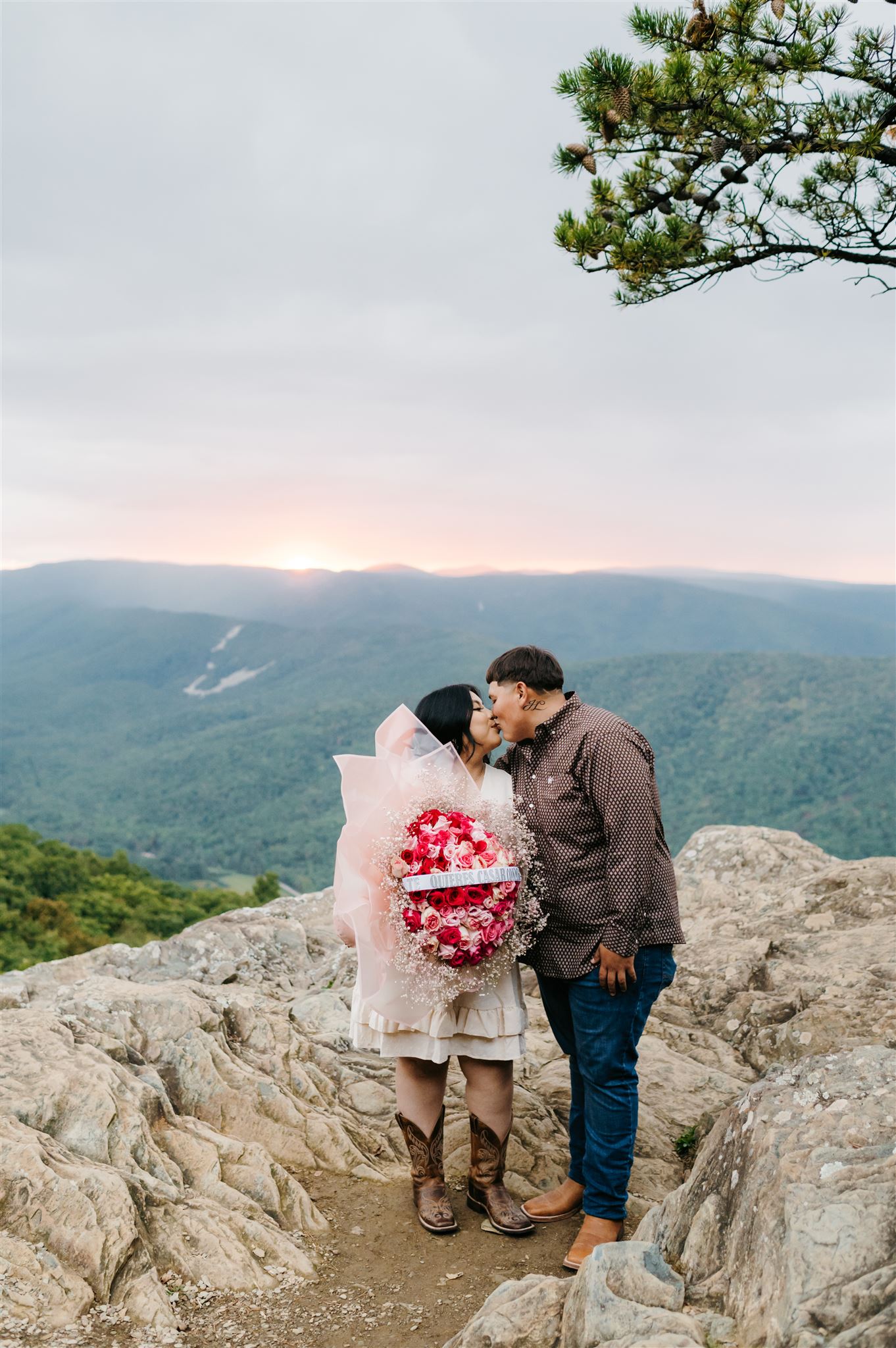 photographers in charlottesville va captures engagement photo with couple kissing while the woman holds a rose bouquet with the Shenandoah mountains in the distance with a light pink sunset