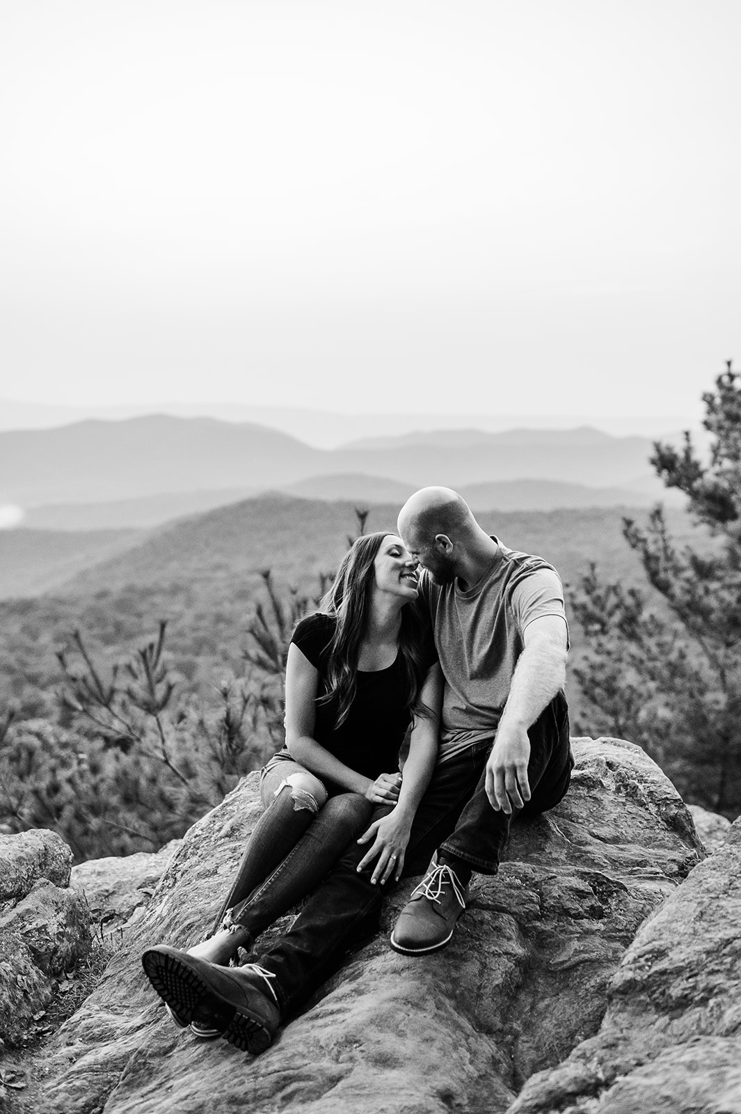 This intimate black-and-white photograph of a couple seated on rocky terrain epitomizes romance and serenity, with the rolling hills of Shenandoah National Park creating a breathtaking backdrop. Their tender connection, captured mid-laughter and gazes filled with warmth, highlights why this location ranks as the best hike in Shenandoah National Park for engagement sessions. The timeless monochrome tone adds depth and emotion to this unforgettable moment.