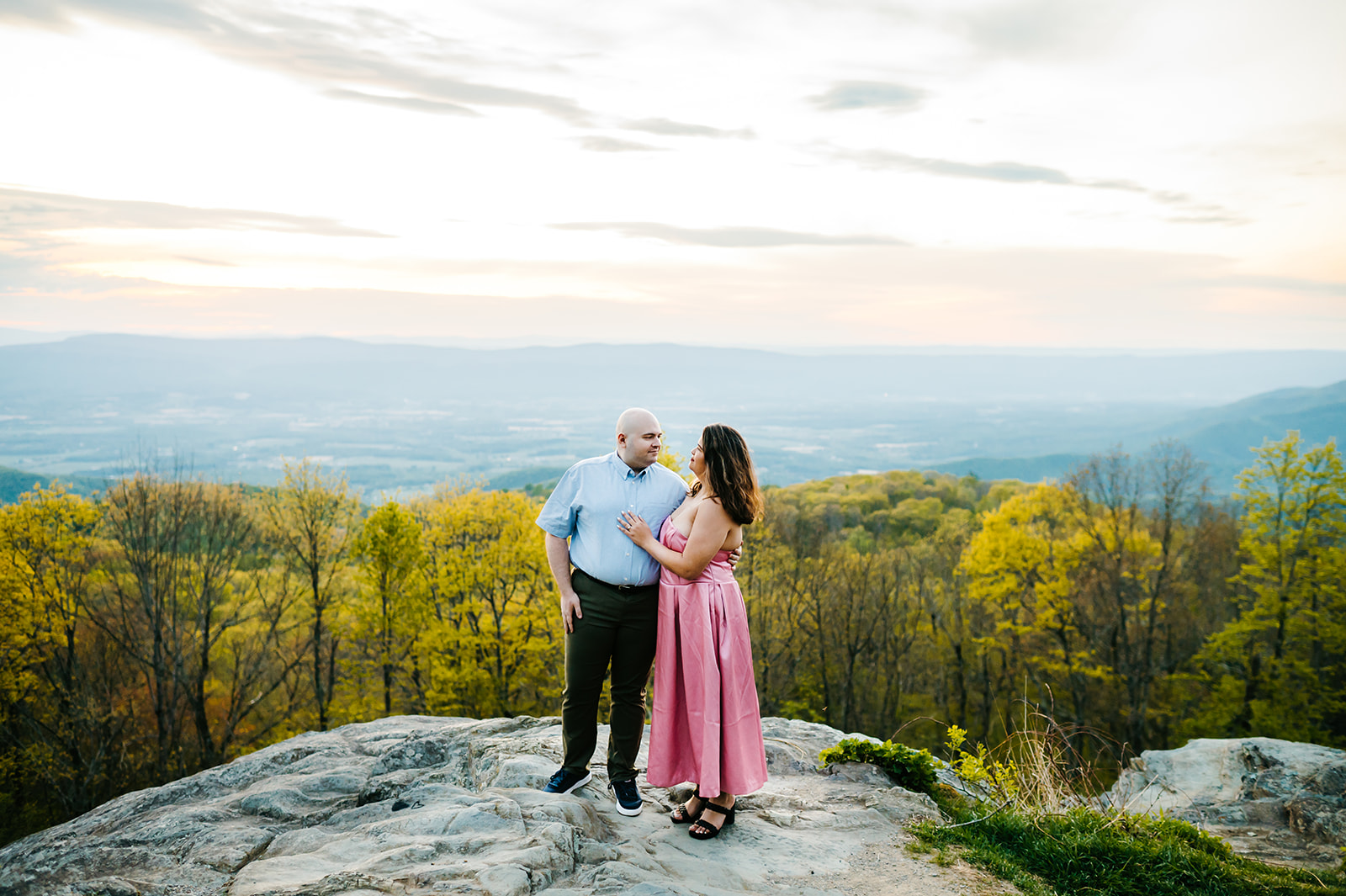 A couple stands together on a rocky overlook with a stunning view of rolling mountains and vibrant spring foliage during their Shenandoah National Park engagement photos. The soft sunset light adds a romantic glow to the scene captured by Charlottesville wedding photographer