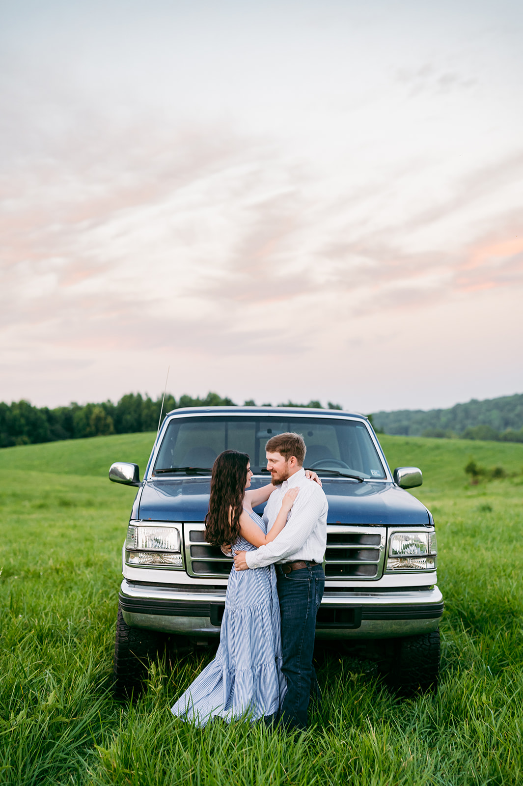 country engagement picture with man and woman holding each other and smiling at sunset in a field with a truck behind them