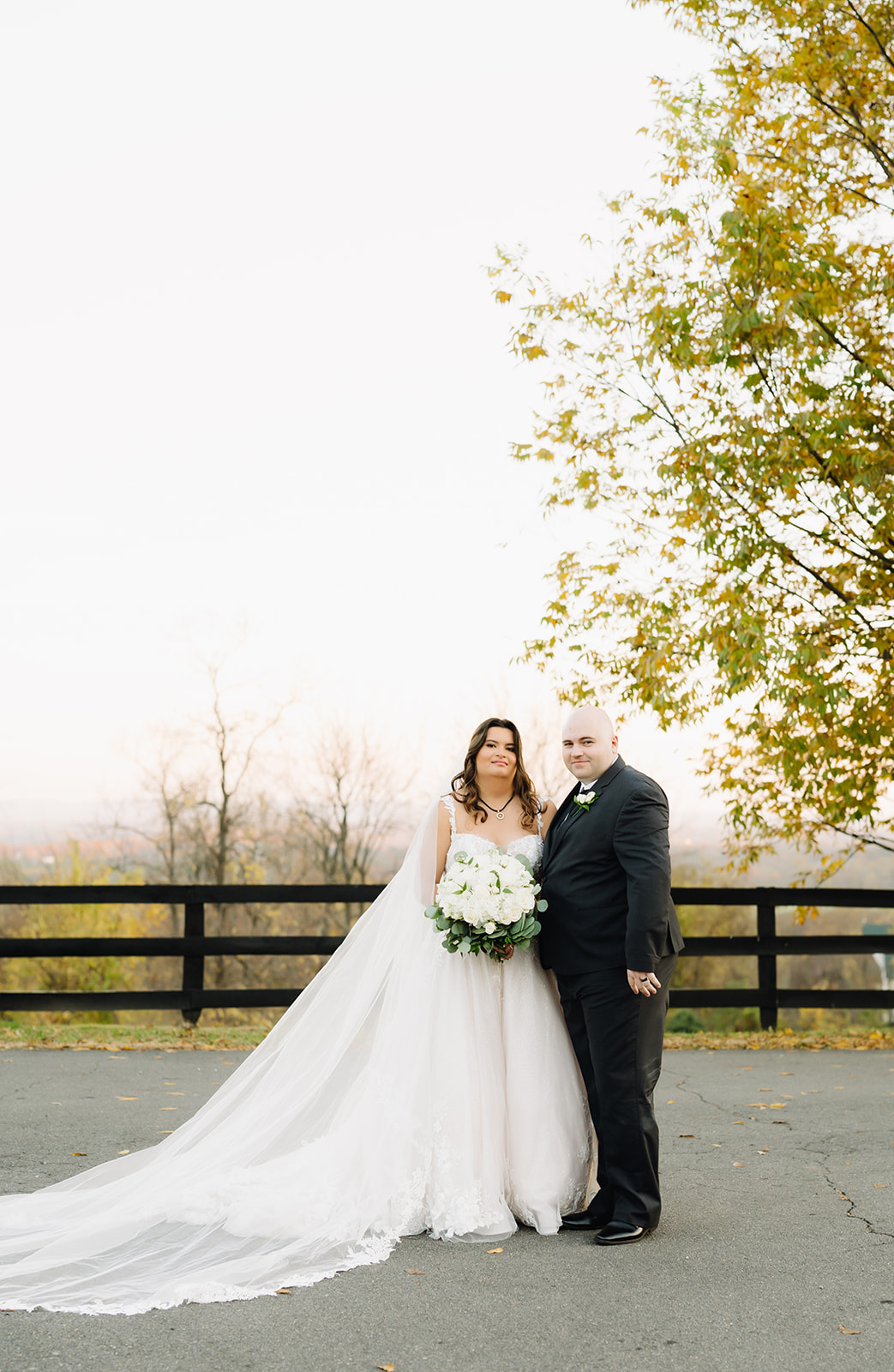 Bride and groom standing together on a paved path at Bluemont Vineyard in Virginia. The bride’s flowing veil cascades behind her, complementing her lace gown and bouquet of white roses and eucalyptus. The groom, in a classic black suit, stands proudly by her side.