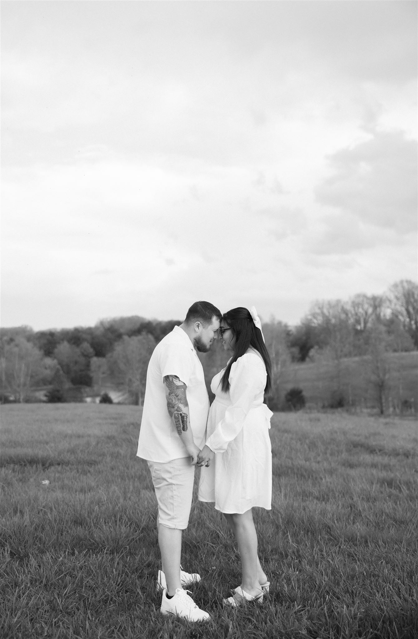 A couple shares an intimate moment during their spring engagement session, standing face-to-face and holding hands in a wide-open grassy field. The black-and-white image highlights their deep connection, with subtle details of the serene countryside and cloudy sky in the background adding a timeless and romantic feel to the scene. photographed by Charlottesville wedding photographer