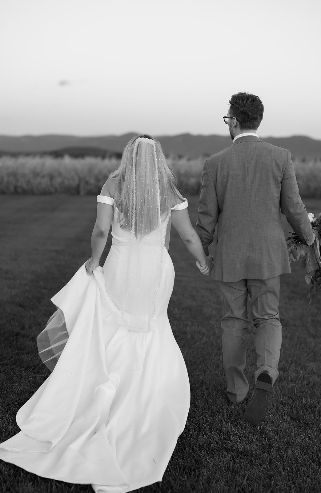 Charlottesville wedding photographer photographs Black and white photograph of a newlywed couple walking hand in hand across a grassy field at Frieden Farms in Shenandoah, with mountains and farmland in the background