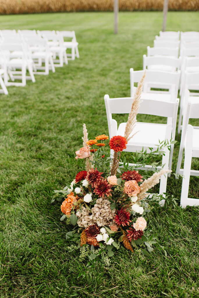 large floral arrangement of orange and red flowers sitting on the ground next to white folding chairs at wedding ceremony at Frieden Farms