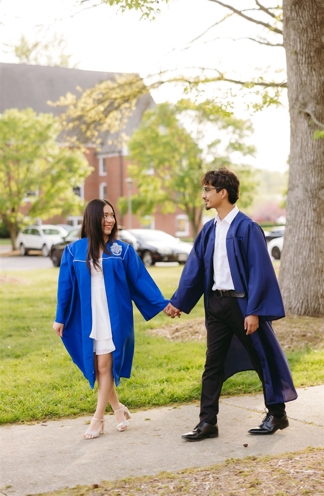 couples photography on college campus with couple holding hands and walking down a path together with buildings and trees in the distance for Couple Graduation Photos