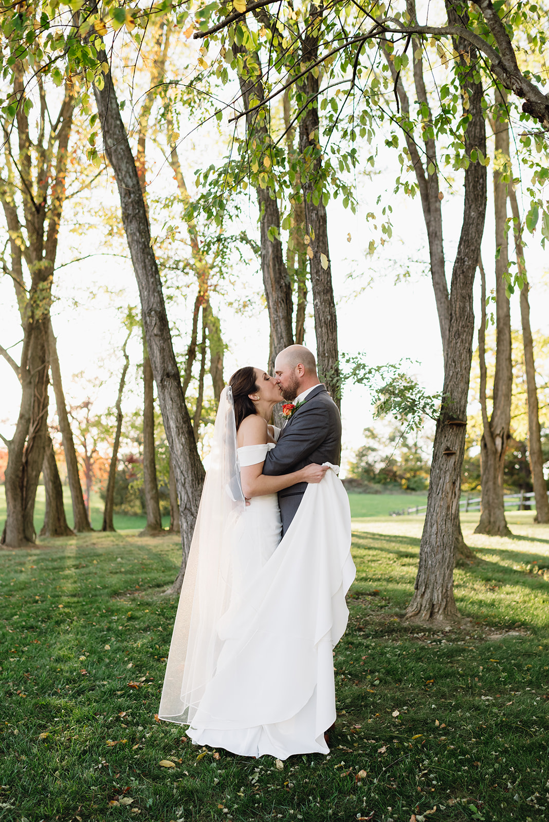 bride and groom share a kiss beneath tall trees at East View Farms and Venue. The groom gently lifts part of the bride’s flowing white gown as they embrace, surrounded by lush greenery and golden sunlight filtering through the leaves photographed by Charlottesville wedding photographer