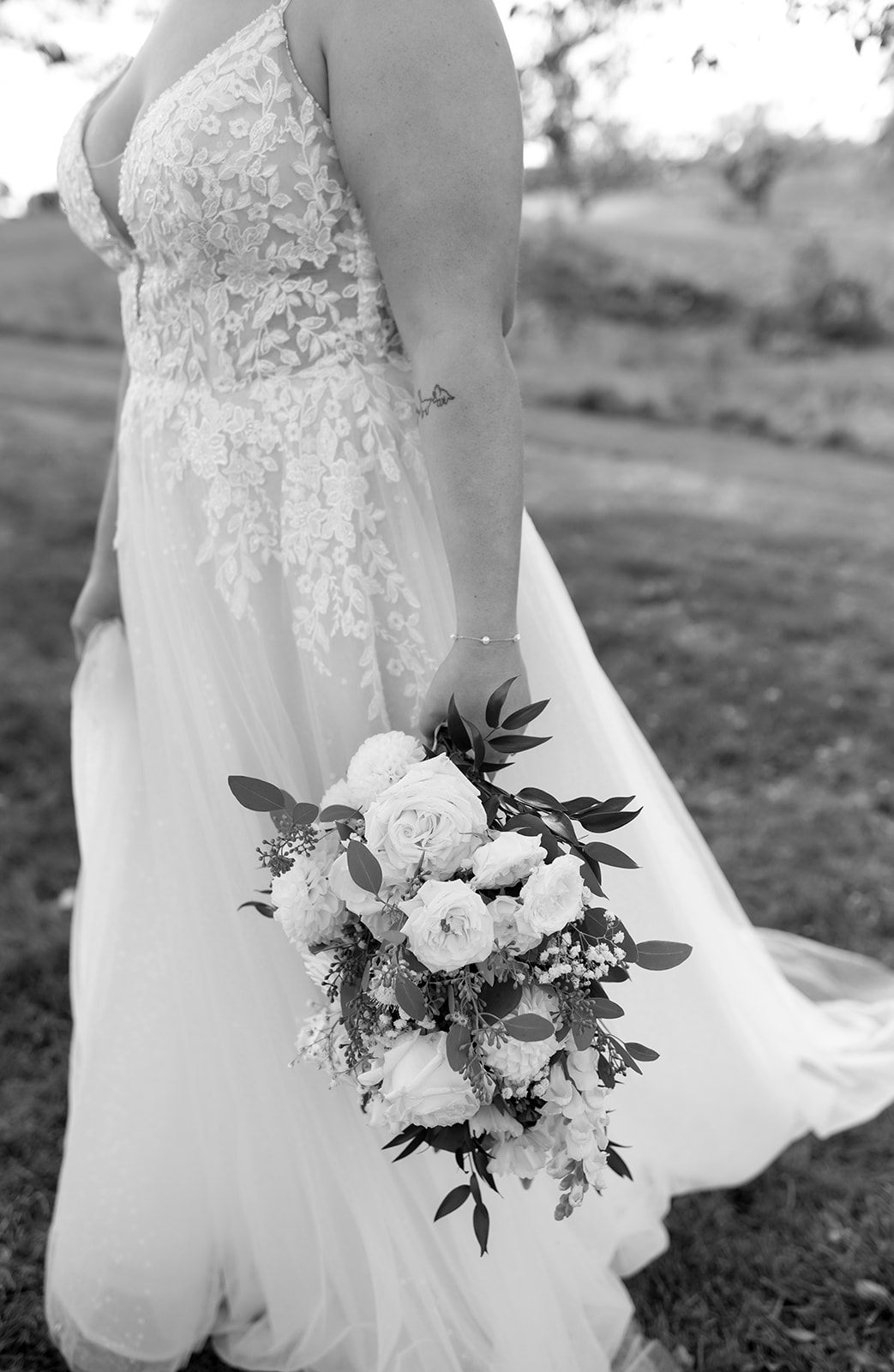 Black and white close-up of a bride at On Sunny Slope Farm Wedding Venue, highlighting the intricate lace details of her wedding gown and the bouquet of roses and greenery she holds by her side