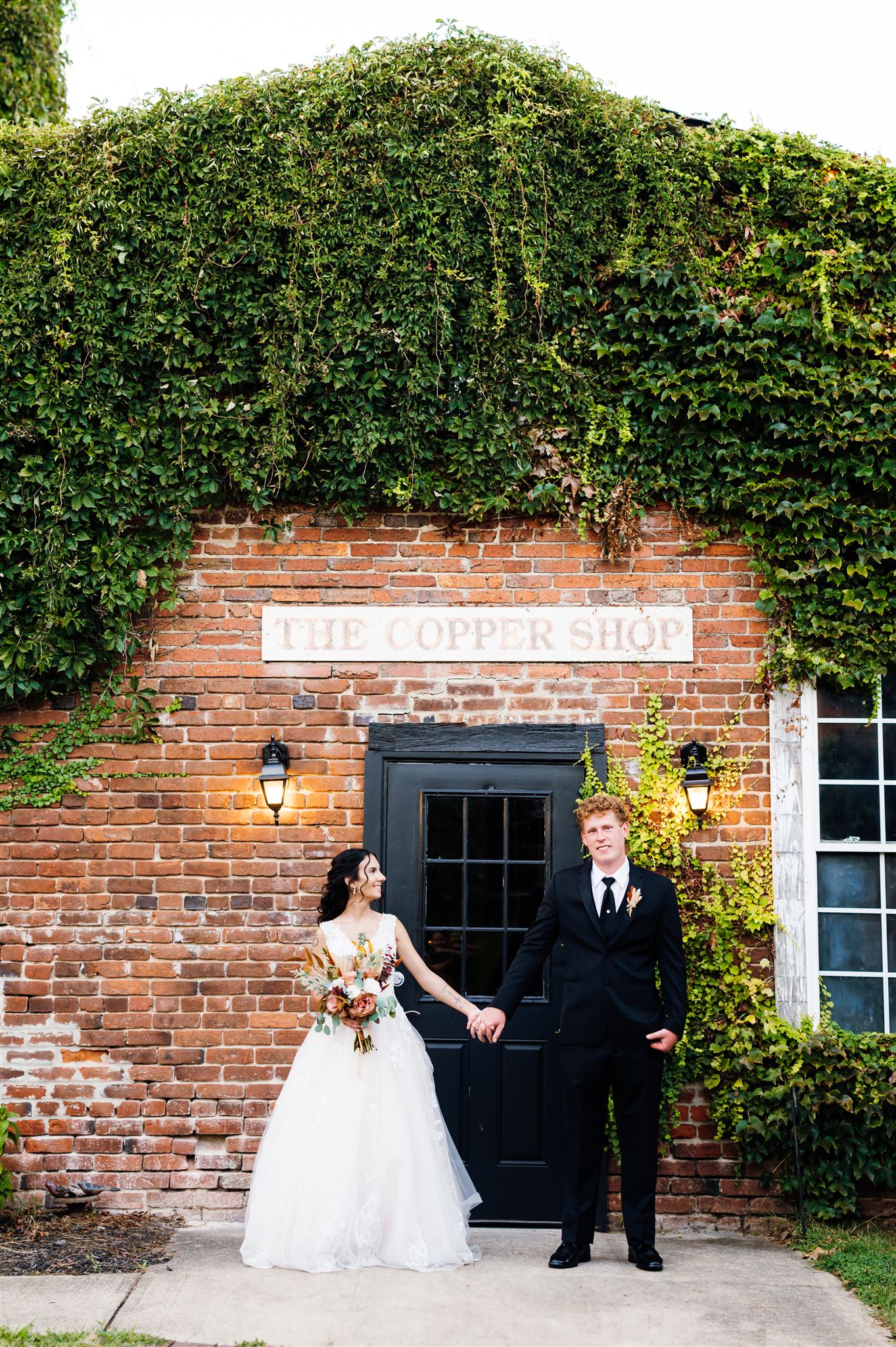 Newlywed couple poses in front of The Copper Shop at the Silk Mill Wedding Venue. The bride, wearing a lace wedding gown and holding a bouquet of roses and greenery, smiles at the groom, who stands in a black suit with a relaxed expression. They hold hands in front of the ivy-covered brick building, framed by vintage lanterns and a black door photographed by Charlottesville wedding photographers